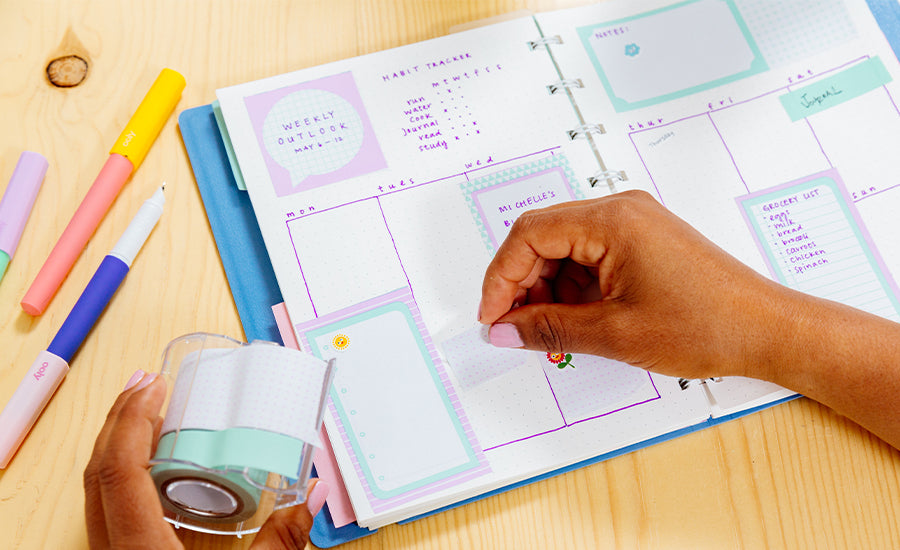 hand putting tape on planner pages on wooden table with colorful pens next to it