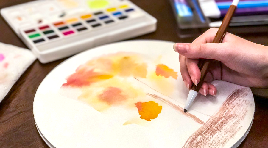 yellow, orange and brown tree being draw on white circular paper with art supplies on brown surface