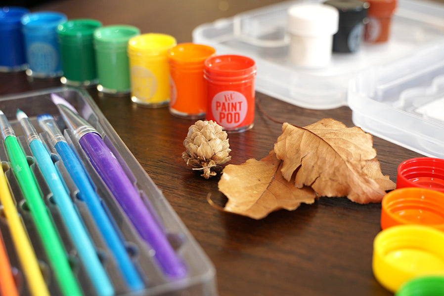 Lil paint pods in rainbow colors next to paint brushes and leaves on a table