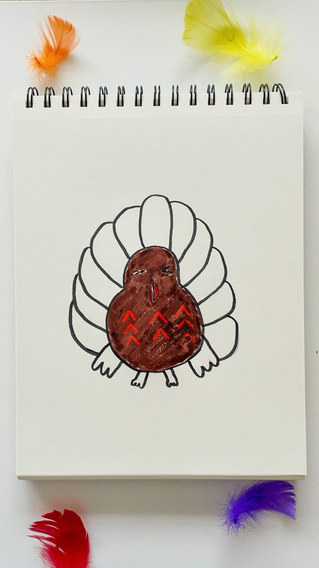 White sketchbook with half colored turkey on white table with colorful feathers