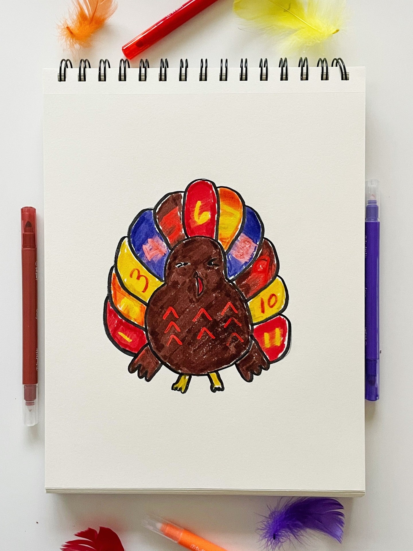 Sketchbook with colored in turkey, markers, and colored feathers on table
