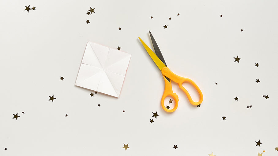 Yellow scissors and white square paper on table with gold stars