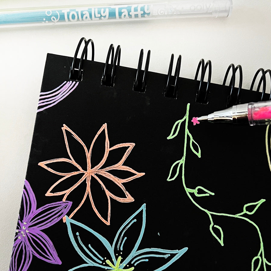 Pink gel pen drawing small flower designs on black sketchbook with totally taffy gel pens in background