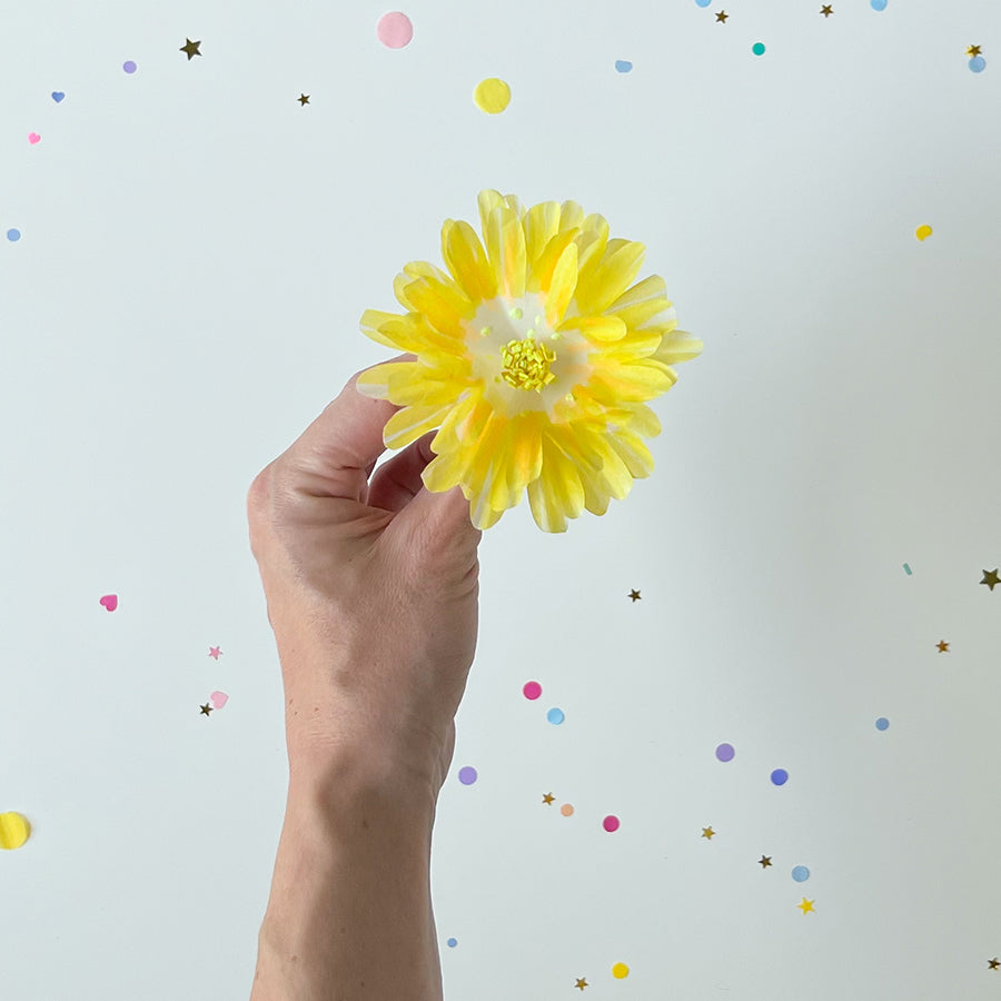 Man's hand holding yellow paper flower with white confetti background