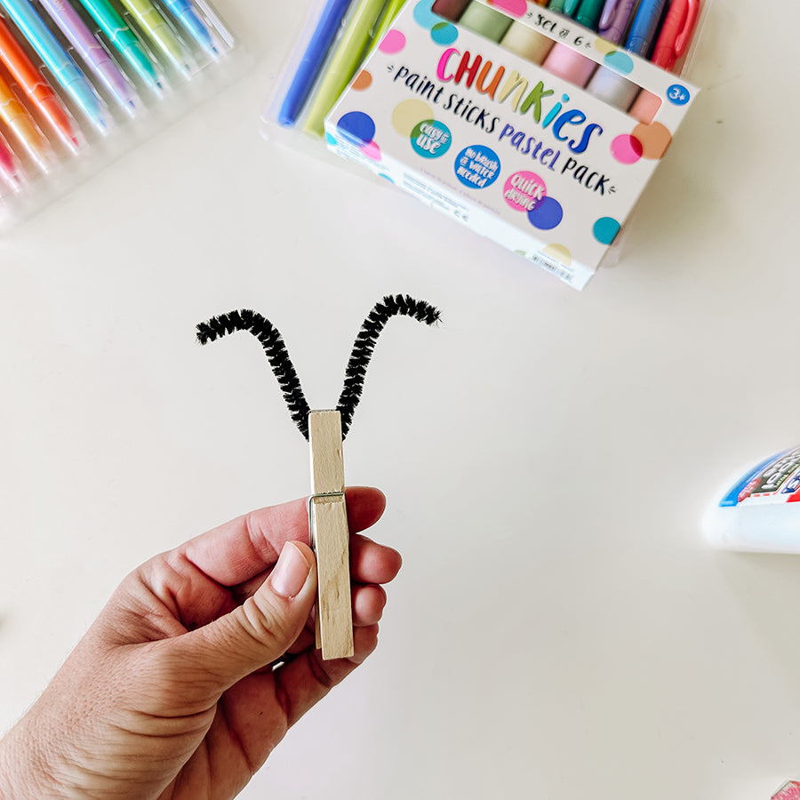 Clothespin holding pipe cleaner in the shape of antenna ears with art supplies in the background