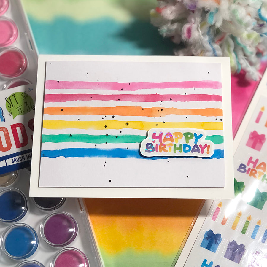 Handmade birthday card with rainbow colors, birthday sticker, and paint pods in background