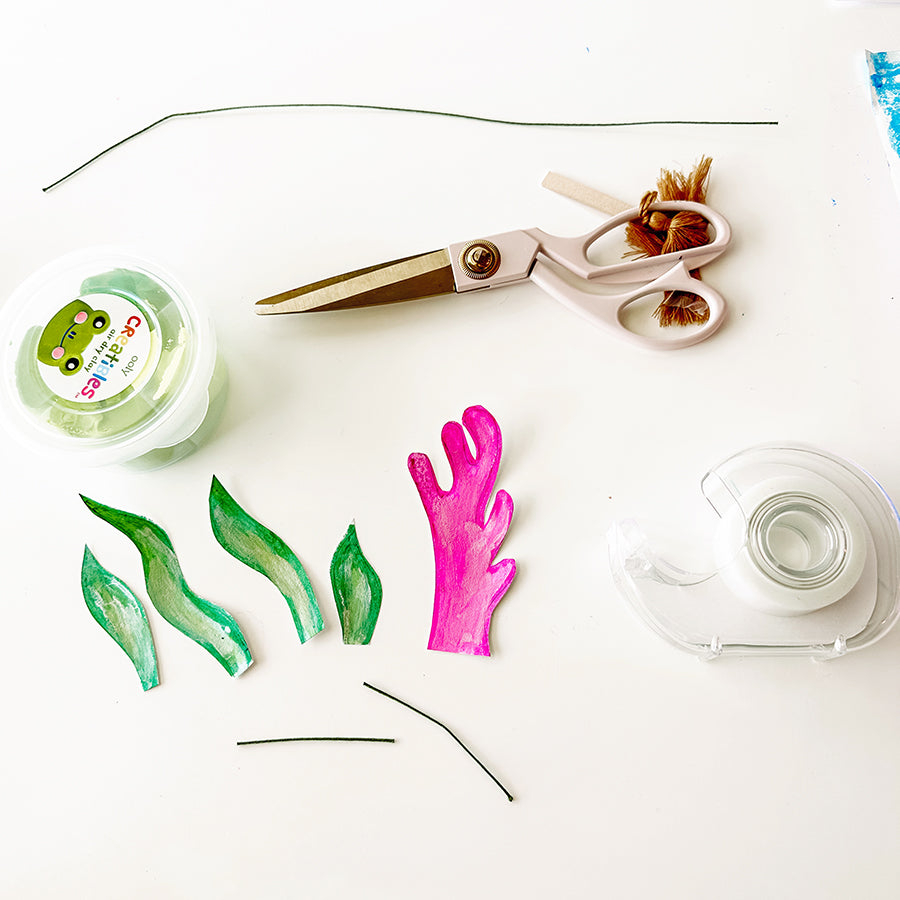 Arts and crafts supplies on table such as scissors, tape, and clay next to cut out shapes of seaweed and coral