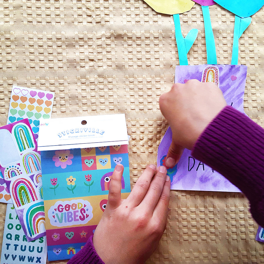 Kid's hands putting stickers on cut out purple vase with more stickers and arts and crafts on the table