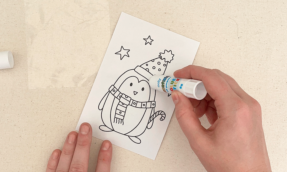 Hand holding ooly glue stick over printable of penguin on table with transparent paper