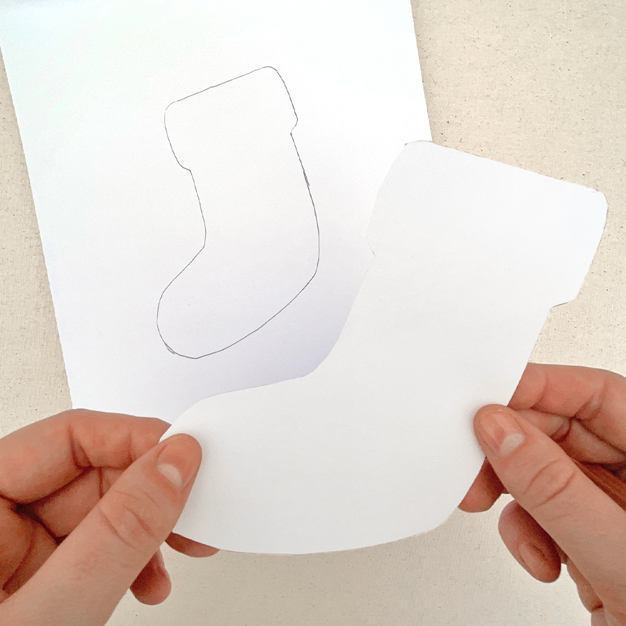 Hand holding the shape of a holiday sock over white sketchpaper with the shape traced on other sketch paper