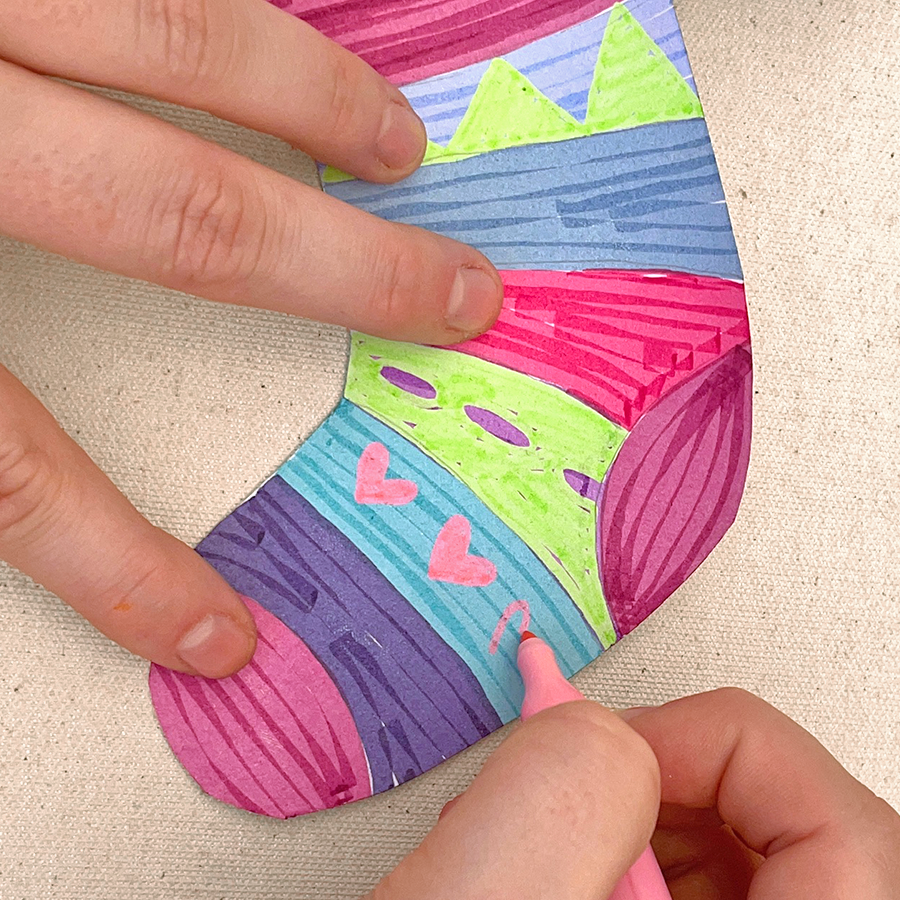 Hand using ooly marker to draw pink hearts on a colorful holiday sock made of paper