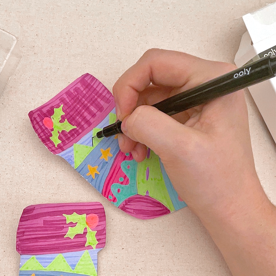 Hand holding ooly ink works markers drawing the outline of triangles on green, purple, and pink Holiday sock